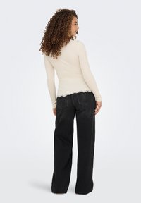 Beige pitkähihaisessa paidassa on röyhelöinen pitsireuna, joka on yhdistetty korkeavyötäröisiin mustiin leveälahkeisiin farkkuihin. Yhdistelmä korostaa rentoa istuvuutta ja tekstuuria.