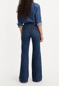 Femme aux cheveux bouclés portant une chemise en denim retroussée et un jean bleu taille haute à jambes larges, debout de dos à la caméra sur un fond uni.