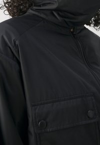 Černá bunda s vysokým límcem a zapínáním na zip, vybavená přední kapsou se zapínáním na druky a hladkou, odolnou texturou látky.