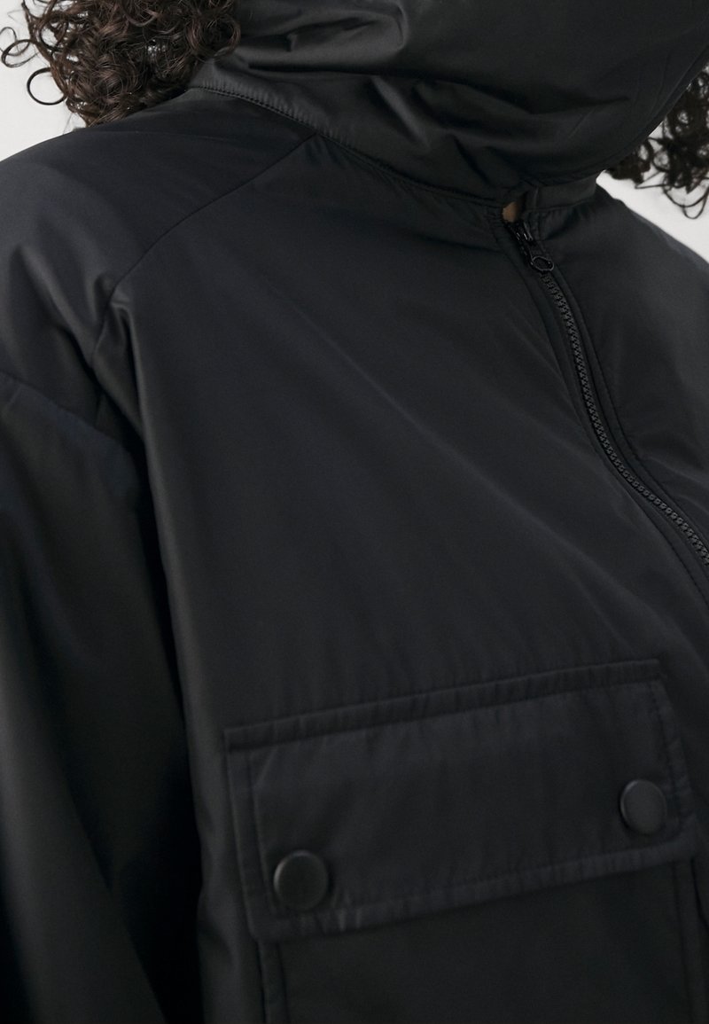Černá bunda s vysokým límcem a zapínáním na zip, vybavená přední kapsou se zapínáním na druky a hladkou, odolnou texturou látky.