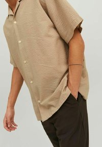 Uomo che indossa una camicia a maniche corte beige testurizzata e pantaloni scuri, con una mano in tasca e un tatuaggio a linea sottile sull'avambraccio.