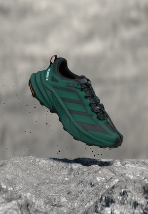 Dunkelgrüner Adidas Terrex Trailrunning-Schuh in der Luft über felsigem Gelände mit umherfliegenden Erdpartikeln.