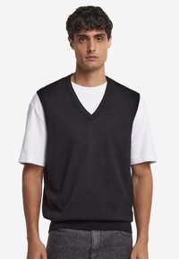 Svart stickad ärmlös tröja med v-ringad design, lager-på-lager över en vit kortärmad skjorta, med ribbad fåll och slät textur.