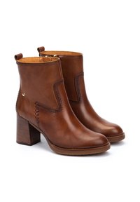 Botas de piel marrón hasta el tobillo con un tacón grueso, que presentan detalles de costura lateral y una pestaña en el tobillo. Superficie suave con punta redondeada.