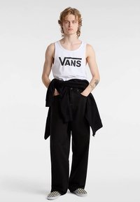 Vit linne med "VANS"-logotyp, svarta stora byxor och en svart tröja knuten i midjan. Skorna har ett svartvitt schackrutigt mönster.