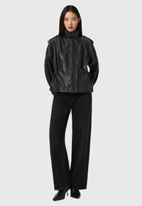 Zwarte leren jas met een hoge kraag, schouderaccenten en een iets losse pasvorm. Gecombineerd met zwarte wijde broek en puntige schoenen.