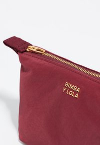 Bolsita de tela burdeos con un zipper dorado y el logo grabado "BIMBA Y LOLA" en un lado. Textura suave y diseño rectangular.