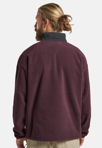 Maroon fleece pullover with a black collar, featuring a relaxed fit and textured surface. Back view shows dropped shoulders and long sleeves.