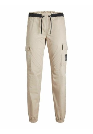 Enfant portant une veste bomber marron, un pantalon cargo beige, des baskets montantes noires avec semelles et lacets blancs, debout sur un fond uni.