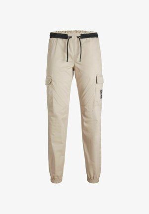 Enfant portant une veste bomber marron, un pantalon cargo beige, des baskets montantes noires avec semelles et lacets blancs, debout sur un fond uni.