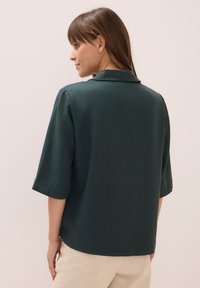 Kvinna med brunt hår klädd i en mörkgrön kortärmad skjorta och beige byxor, stående mot en ljus och enkel bakgrund, med sidan mot kameran.