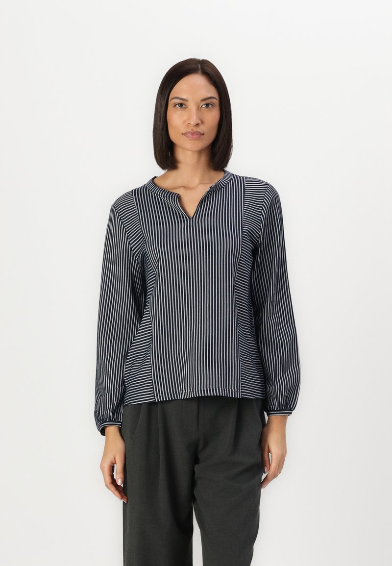 Striped blouse with a V-neckline, featuring long sleeves and gathered cuffs, in navy blue and white. Worn with grey trousers.