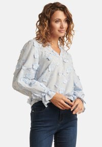 Light blue blouse with floral embroidery, v-neck, and ruffled cuffs. Paired with dark blue jeans. Soft fabric and textured detailing.