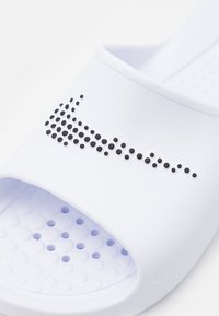 White slide sandal with a textured upper, featuring a prominent black dotted Nike logo. The sole has perforations for breathability.