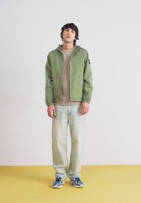 Green lightweight bomber jacket, beige crewneck shirt, faded blue jeans, and blue sneakers. Simple design, relaxed fit, and textured fabric.