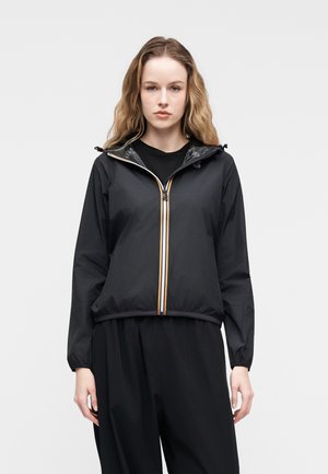 Giovane donna con capelli mossi che indossa una giacca nera con cappuccio e una cerniera a righe, in piedi davanti a uno sfondo chiaro e uniforme.