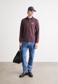 Cămașă polo lungă bordo cu guler albastru închis și emblemă, purtată cu blugi deschiși la culoare și cizme negre până la gleznă. Ține în mână o geantă texturată neagră.