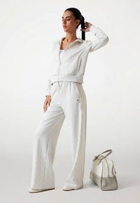 Tuta bianca con giacca con zip e pantaloni a gamba larga, caratterizzata da un motivo sottile e tessuto strutturato. Accompagnata da una borsa leggera.