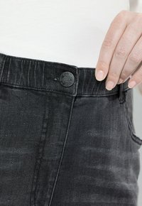 Ruka upravující pás kalhot z černého denimového materiálu s kovovým knoflíkem, na sobě bílý top, detailní záběr na pas a ruku.