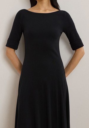Frau trägt ein eng anliegendes schwarzes Kleid mit hochgekrempelten Ärmeln bis zum Ellbogen und steht mit den Händen hinter dem Rücken vor einem einfarbigen Hintergrund.