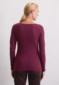 Langarm, tailliertes Hemd in tiefem Lila, mit weitem Ausschnitt und strukturiertem Stoff. Glatte Nähte mit leichtem Schwung am Saum.