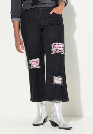 Jeans amplios de mezclilla negros con parches desgastados que revelan una tela a rayas en rosa y blanco, combinados con botines plateados. Tejido de mezclilla texturizado.
