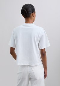 Vrouw met een lage bun die een eenvoudige witte T-shirt met korte mouwen en witte broek draagt, gezien van achteren tegen een effen achtergrond.