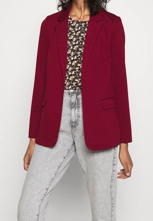 Femme portant un blazer rouge foncé sur un top noir fleuri associé à un jean taille haute gris clair sur un fond clair.