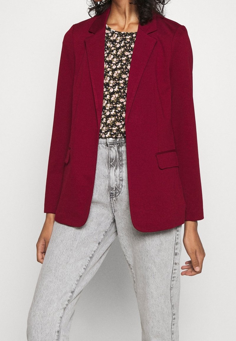 Femme portant un blazer rouge foncé sur un top noir fleuri associé à un jean taille haute gris clair sur un fond clair.