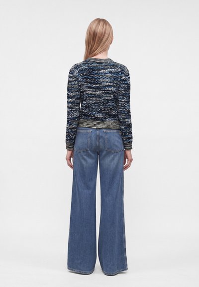 Femme aux longs cheveux blonds portant un pull bleu à motifs et un jean bleu à jambes larges, debout de dos devant un fond uni.