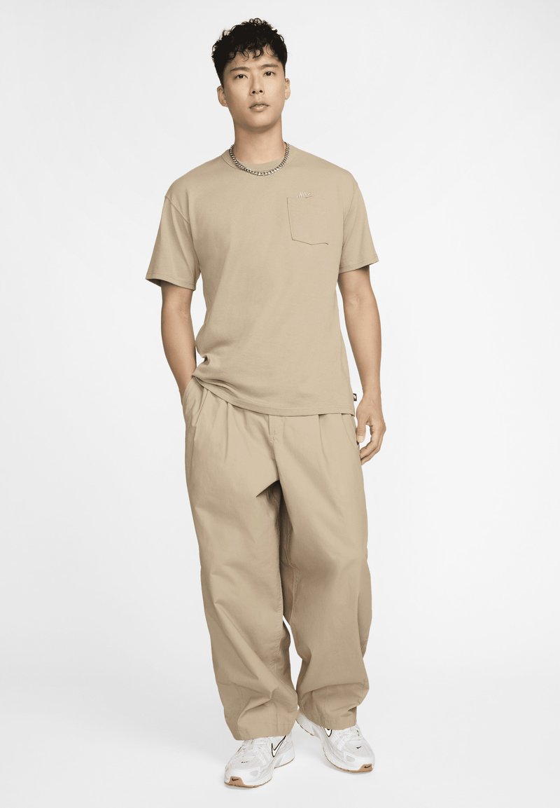 Camiseta de algodón beige con un bolsillo en el pecho, combinada con pantalones beige holgados y zapatillas blancas. El atuendo presenta un diseño relajado y casual.