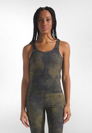 Donna con lunghe trecce che indossa una canotta verde e nera con motivo senza maniche e leggings aderenti abbinati, in piedi davanti a uno sfondo bianco semplice.