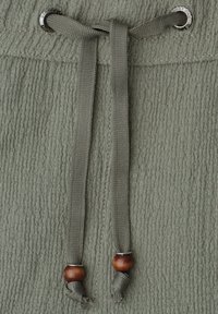 Gros plan d’un tissu texturé vert olive avec des cordons assortis enfilés dans des œillets métalliques, chacun se terminant par une perle en bois.