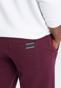 Burgunderfarbene Sweatpants mit einer strukturierten Oberfläche, mit einem schwarzen Metall-Logoetikett auf der Rückentasche und einem glatten weißen Shirt darüber.