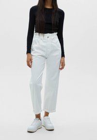 Weiße hochgeschnittene Jeans mit lockerer Passform, mit umgeschlagenem Saum, zwei Fronttaschen und kombiniert mit weißen Sneakers und einem schwarzen Langarmoberteil.