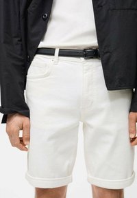 Weiße Denim-Shorts mit umgeschlagenen Säumen, ausgestattet mit einem Knopf- und Reißverschluss, kombiniert mit einem schwarzen Gürtel und einer schwarzen Jacke.