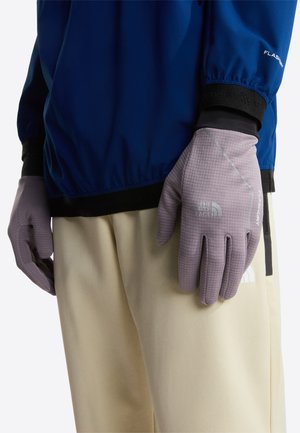 Gants gris clair avec une prise en main texturée, arborant un logo et des accents réfléchissants, portés par une personne en veste bleue et pantalon beige.
