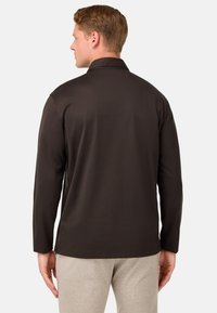 Garais piedurknu krekls tumši brūnā krāsā ar apkakli, gludu tekstūru, sānu šķēlumiem apakšmalā un valkāts pār gaiši pelēkiem sporta biksēm.