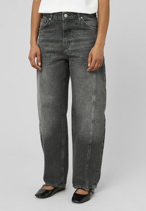 Personne portant un jean gris foncé ample à taille haute avec un haut blanc et des ballerines noires devant un fond uni.