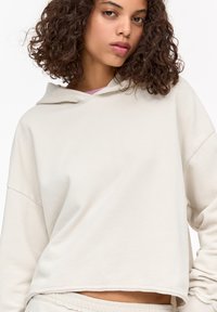 Lichtbeige cropped hoodie met een trekkoordkap, verlaagde schouders en een zachte textuur; voorzien van ruwe zoomdetails en een minimalistisch ontwerp.