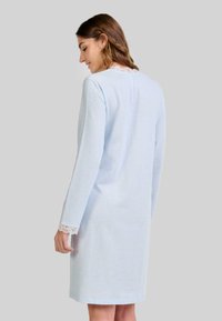 Mujer con un vestido de mangas largas de un azul claro con encaje blanco en el cuello y los puños, vista desde atrás contra un fondo sencillo.