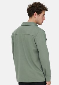 Mann mit lockigem Haar, der ein hellgrünes Langarmshirt trägt, von hinten gezeigt und leicht nach rechts gedreht.