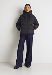 Black padded jacket with a high collar and drawstring hood, featuring a zip closure and side pockets, paired with wide-leg dark denim trousers.