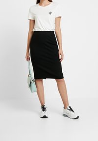 Kvinna som bär en vit t-shirt med ormlogga, svart knälång kjol, vita sneakers och håller en ljusblå handväska vid sin sida.