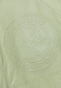 Hellgrüne Lederoberfläche mit einem geprägten runden Logo, das einen Baum und den Text „ELKVIS EST. 2013“ zeigt.