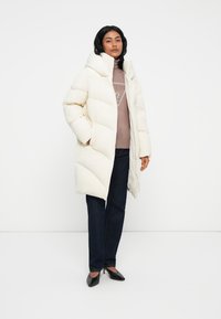 Žena v dlouhém bílém péřovém kabátu, svetru v barvě mauve s logem, tmavých jeans a černých špičatých lodičkách na bílém pozadí.