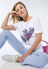 Witte T-shirt met korte mouwen, versierd met een kleurrijke hartafdruk in paarse en blauwe patronen. Gecombineerd met lichtblauwe broek en witte sneakers.