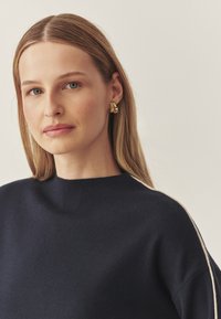 Marineblauer gestrickter Pullover mit hohem Kragen und glatter Textur, ergänzt durch cremefarbene Seitenstreifen und goldene strukturierte Ohrringe.