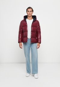 Young man wearing burgundy puffer jacket over white shirt, light blue wide-leg jeans, and white sneakers standing against plain background.