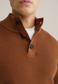 Ruda megzta džemperis su aukštu apykakle, turintis tekstūruotą raštą ir tris juodus mygtukus prie kaklo. Dėvimas ant balto marškinėlio.